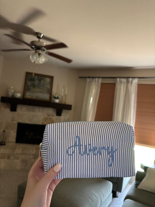 A hand holding a small zippered pouch with blue and white stripes and the name 'Avery' embroidered in blue script, shown in a home interior setting.