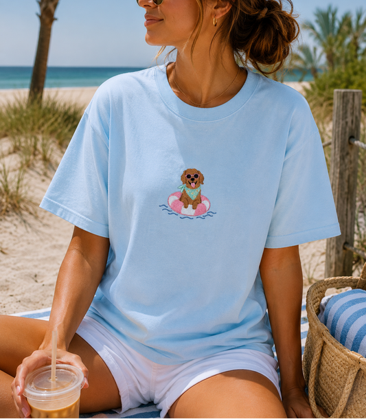 A woman sitting on a beach chair at the beach wearing a light blue crewneck t-shirt with a cartoon dog graphic on the chest, paired with white shorts.