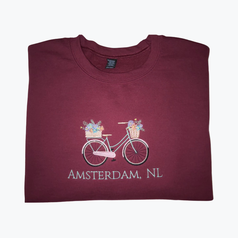 A folded maroon crewneck t-shirt featuring a graphic of a bicycle with flower baskets and the text 'AMSTERDAM, NL' printed below it, displayed on a clean white background.