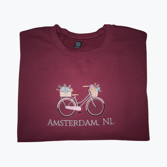A folded maroon crewneck t-shirt featuring a graphic of a bicycle with flower baskets and the text 'AMSTERDAM, NL' printed below it, displayed on a clean white background.