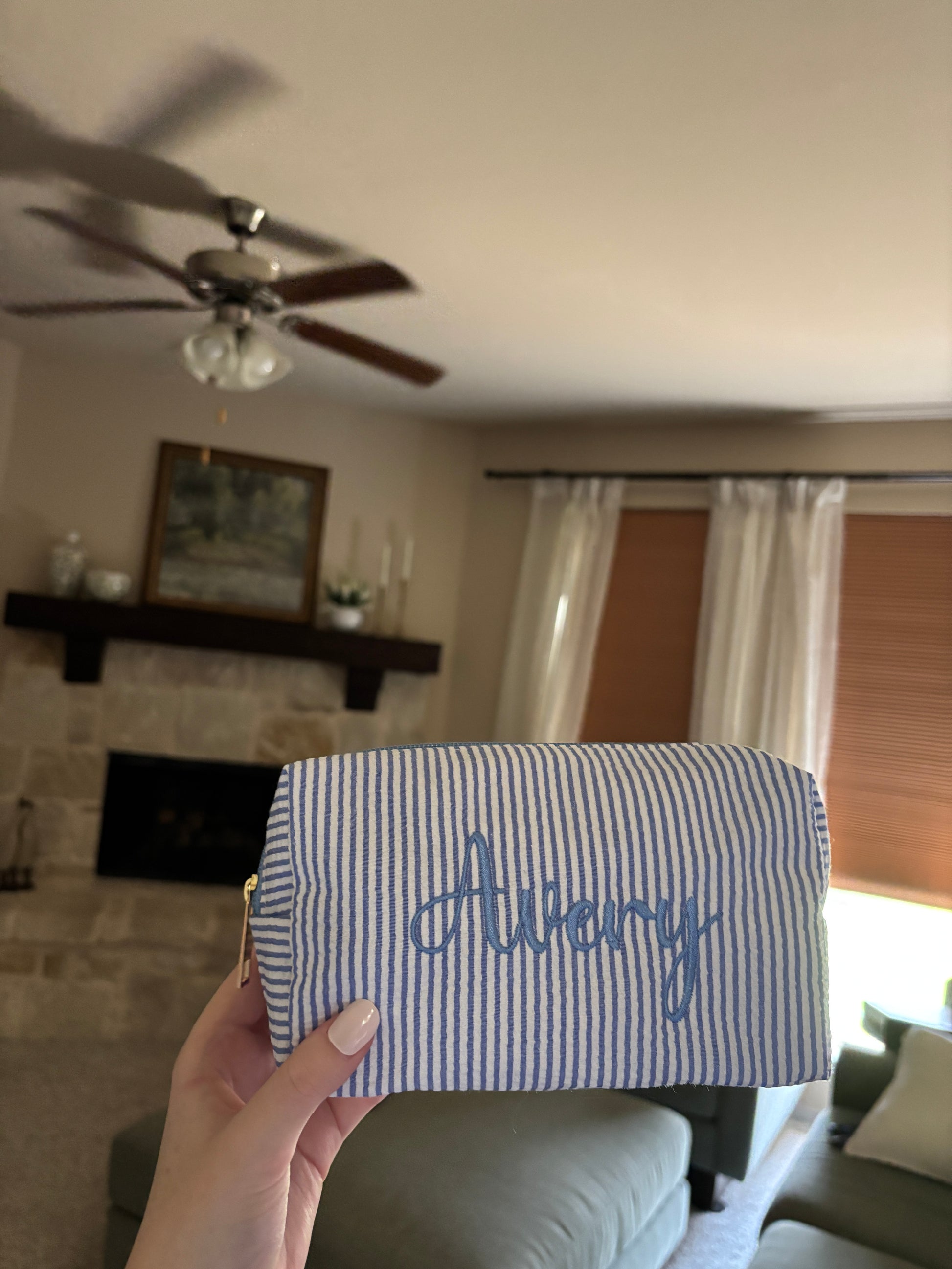 A hand holding a small zippered pouch with blue and white stripes and the name 'Avery' embroidered in blue script, shown in a home interior setting.