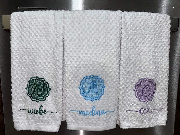 Three white textured dishtowels with embroidered monograms and names hanging on a dishwasher