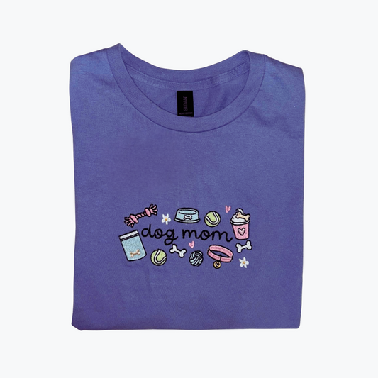 Folded purple t-shirt with 'dog mom' script style text and illustrations (dog toys, pupiccino, collar, bones, treats) on a white background. 