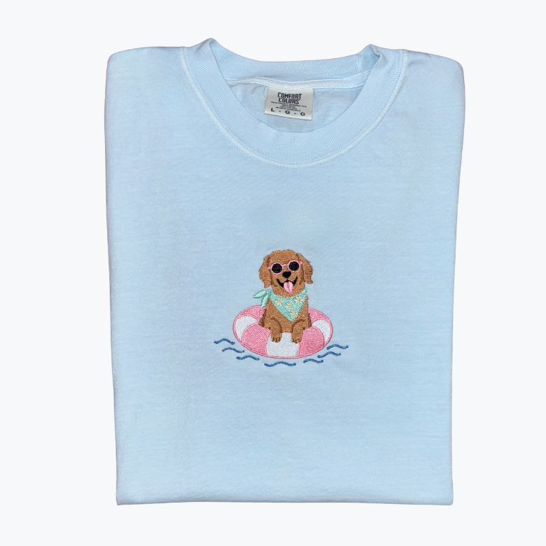 A light blue folded crewneck t-shirt featuring a cartoon graphic of a brown dog wearing sunglasses and a green bandana, floating on a pink and white inner tube with blue water lines.