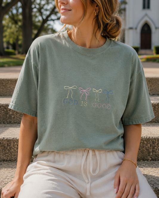 A woman wearing a sage green graphic t-shirt with embroidered bows and the text 'GOD IS GOOD' sitting on outdoor stone steps.