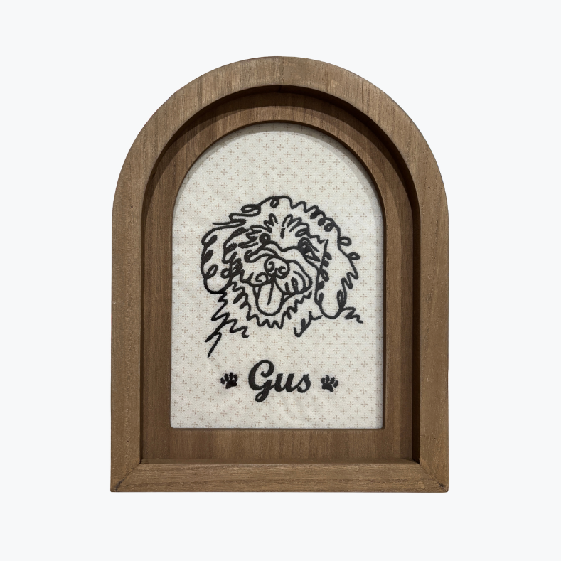 An arched wooden picture frame with a black line art illustration of a Bernedoodle face and the name 'Gus' with paw prints, set against a light textured background.
