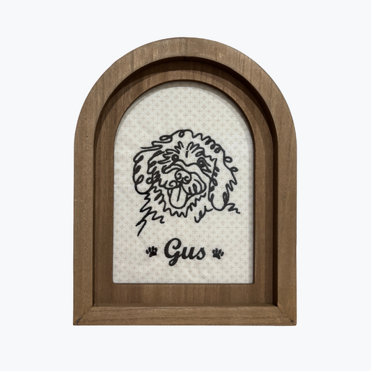 An arched wooden picture frame with a black line art illustration of a Bernedoodle face and the name 'Gus' with paw prints, set against a light textured background.
