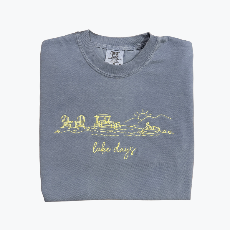 A folded slate blue t-shirt with a yellow line art graphic of a lakefront scene and the text 'lake days' on the front, displayed against a clean white background.