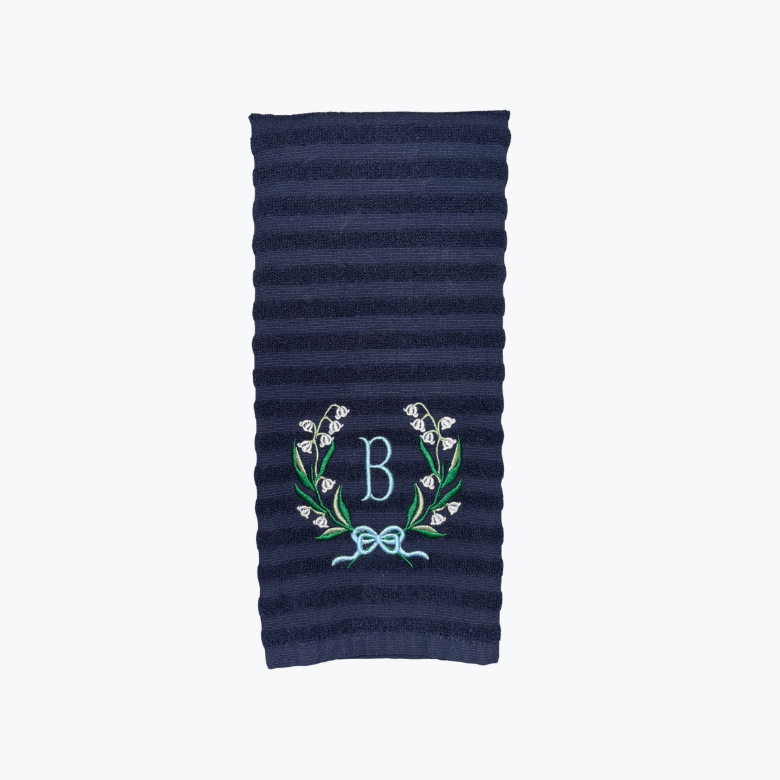 Navy blue dishtowel folded over with a lily of the valley in a circular frame, with a blue "B" initial and light blue bow in the middle of the design.