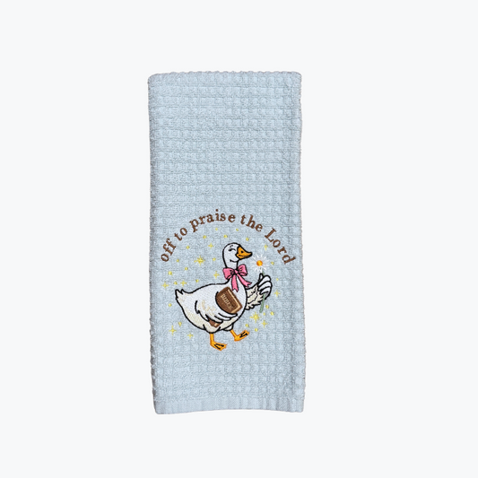 A light blue waffle-weave kitchen towel featuring a cartoon goose wearing a pink bow and the text 'God to praise the Lord' in brown, shown against a plain white background.