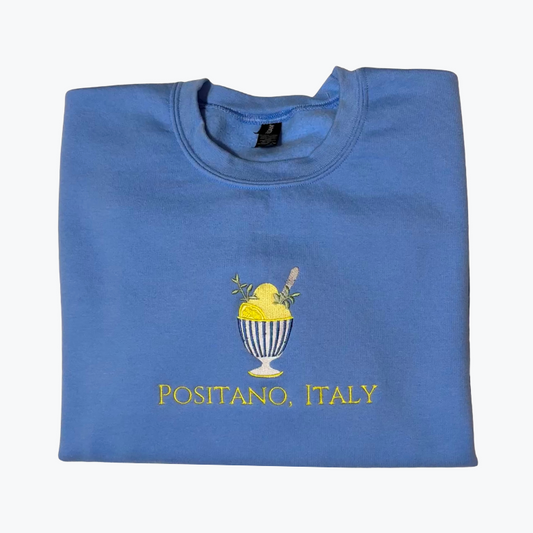 A folded blue crewneck t-shirt featuring a graphic of a yellow lemon gelato sundae in a striped glass and the text 'POSITANO, ITALY' printed below in yellow.