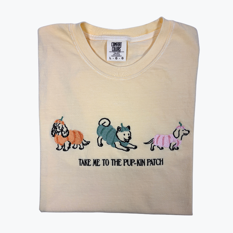 A folded butter-colored t-shirt with embroidered cartoon dogs and the text 'TAKE ME TO THE PUP-KIN PATCH' on the front, displayed against a plain white background.