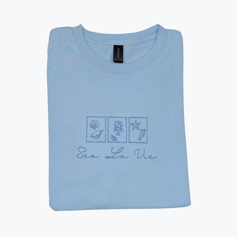 Folded light blue t-shirt with 'Sea La Vie' design of scalloped stamps with ocean themed icons inside of each stamp (3 total)