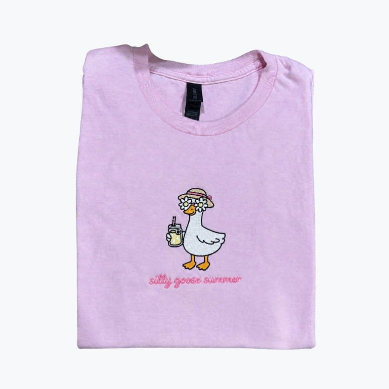 A folded light purple t-shirt with a cartoon graphic of a white goose wearing a straw hat and holding a drink, with the text 'silly goose summer' printed below it, on a plain white background.