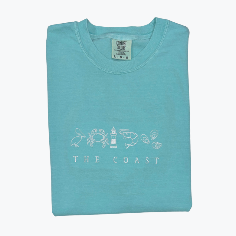 Folded turquoise t-shirt with coastal design and "The Coast" text
