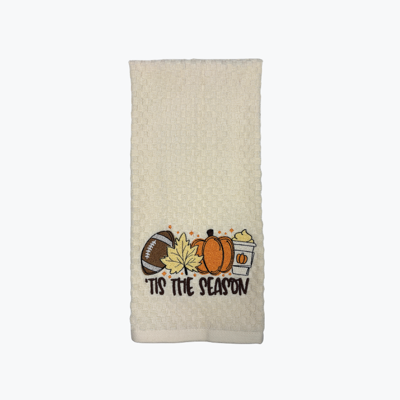 An Ivory-colored kitchen towel featuring a fall-themed graphic with a football, pumpkin, maple leaf, drink, and the text 'TIs THE SEASON' 