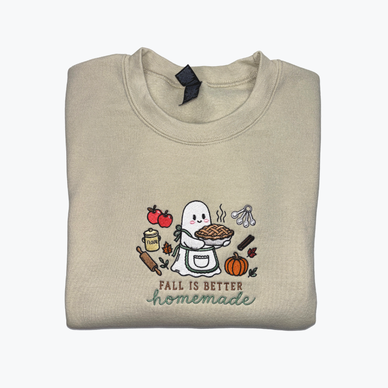 A folded light beige crewneck sweatshirt featuring a cute cartoon graphic of a ghost baking a pie with autumn-themed illustrations and the text 'FALL IS BETTER homemade'.