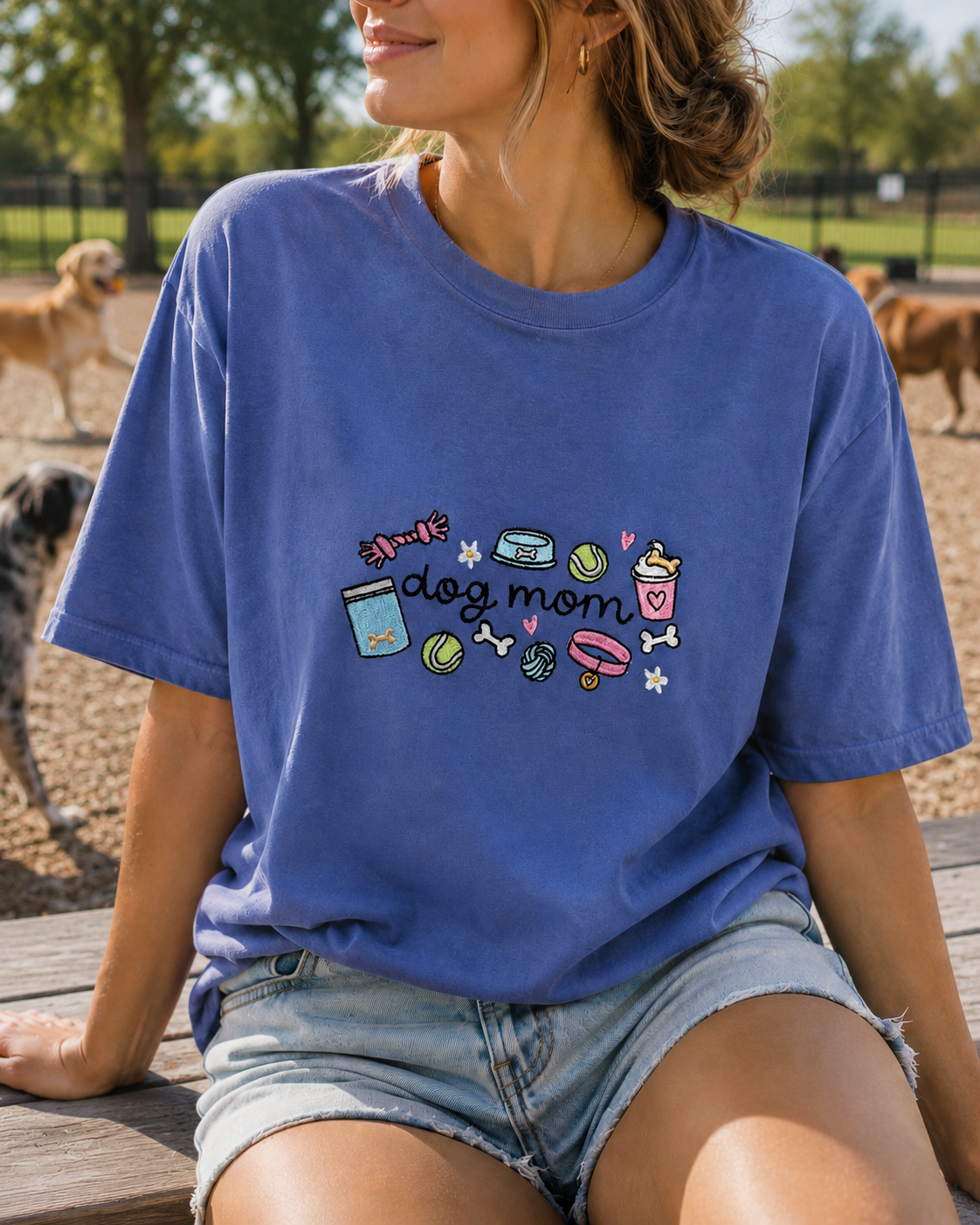 A woman wearing a blue graphic t-shirt with a 'dog mom' design, sitting on a wooden bench in an outdoor dog park.