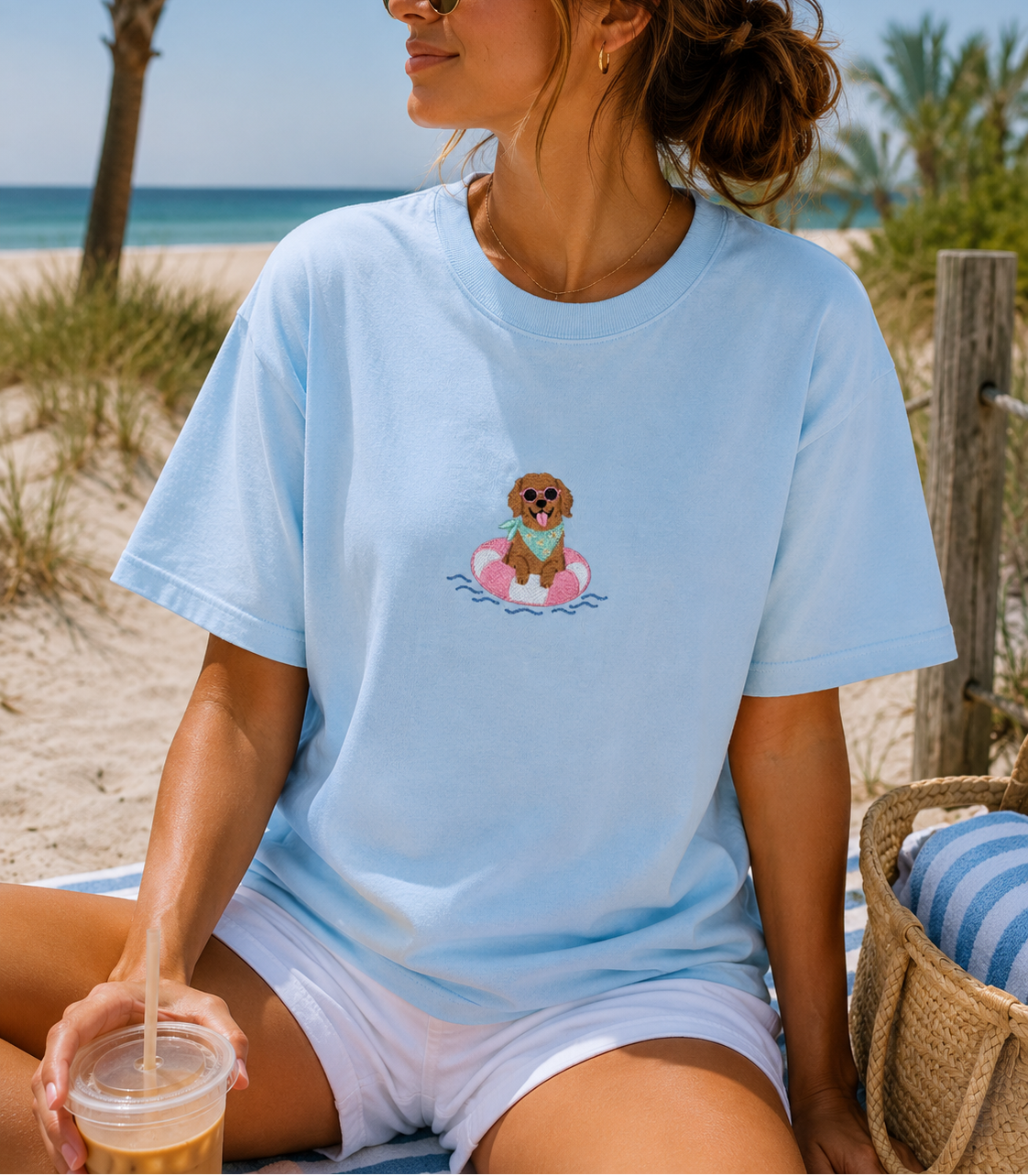 A woman sitting on a beach chair at the beach wearing a light blue crewneck t-shirt with a cartoon dog graphic on the chest, paired with white shorts.