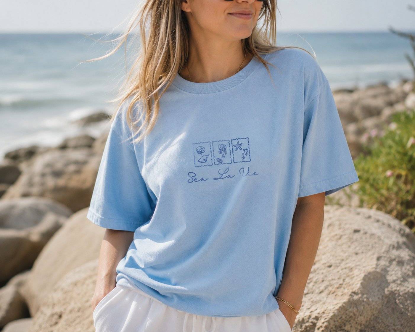 A woman wearing a light blue crewneck t-shirt with a 'Sea Life' logo graphic on the chest, standing on rocks with the ocean in the background.