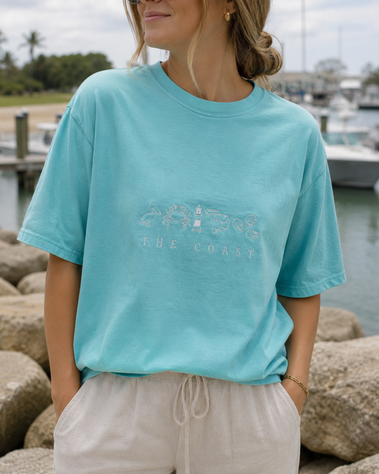 A woman wearing a light turquoise crewneck t-shirt with a white 'THE COAST' graphic on the front, paired with light beige drawstring pants, standing outdoors near rocks and water.
